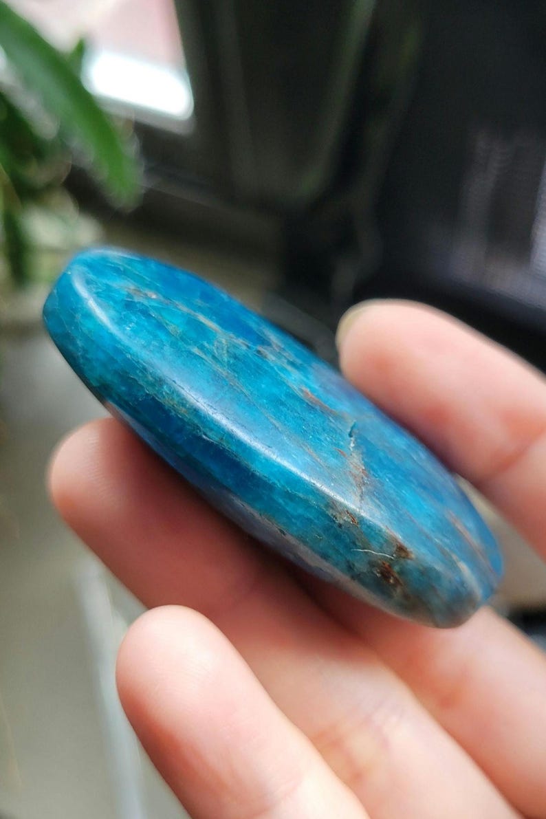 May include: A polished, oval-shaped blue apatite gemstone. The stone has a smooth surface with natural variations in colour and veining. The gemstone is held in a hand, with a blurred background.