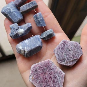 May include: A collection of raw gemstones in shades of blue and pink. The blue stones are rectangular and irregular, while the pink stones are hexagonal. The gems are held in a hand, showcasing their natural textures and colors.