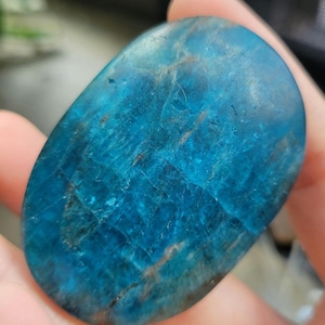 May include: An oval-shaped blue apatite gemstone with a polished surface. The stone has a vibrant, translucent blue colour with brown and white veining. The gemstone is held in a hand, showcasing its natural beauty and unique patterns.