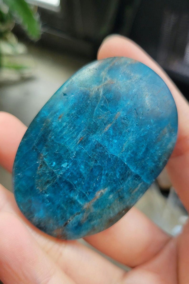 May include: An oval-shaped blue apatite gemstone with a polished surface. The stone has a vibrant, translucent blue colour with brown and white veining. The gemstone is held in a hand, showcasing its natural beauty and unique patterns.