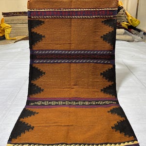 May include: A rectangular, hand-woven rug with a brown central field and black geometric borders. The rug features bands of red, purple, and blue accents. The rug is made of natural fibers and is suitable for a hallway or entryway.