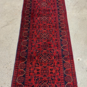 May include: A long, rectangular red rug with an intricate geometric pattern. The rug features a central field of red with navy blue and gold accents. The design includes repeating floral and diamond shapes, creating a traditional aesthetic.