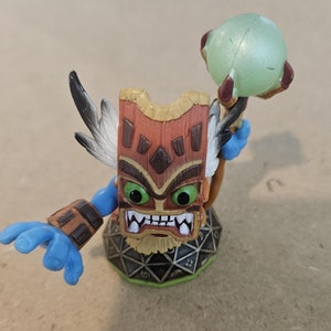 May include: A brown and blue tiki-themed action figure holding a green orb. The figure is standing on a black and green base with a geometric pattern.