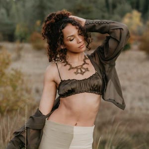 Puede incluir: Una mujer con un top corto verde oliva oscuro y una chaqueta transparente a juego. También lleva un collar único de color bronce. El conjunto se combina con una falda de dos tonos en tonos beige y blanco. El fondo es una escena borrosa al aire libre.