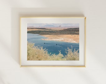May include: A framed landscape print showing a blue lake with boats, set against arid terrain and mountains under a bright sky. The print is presented in a gold frame.