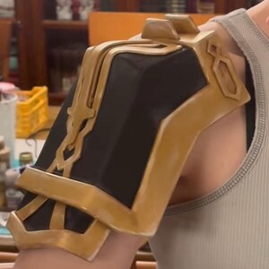 May include: A gold and black shoulder armor piece with intricate details. The armor is made of a hard plastic material and has a textured surface.