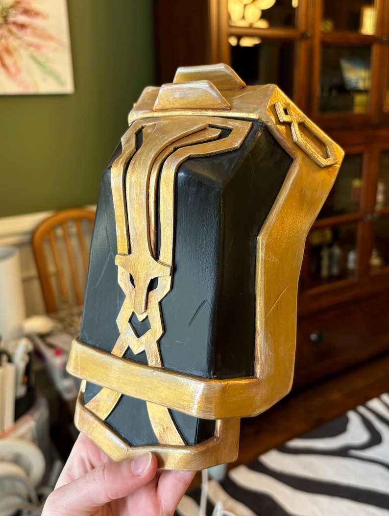 May include: A black and gold decorative piece with intricate details. The piece is made of a hard material and has a smooth, polished finish.