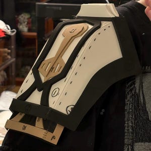 May include: A white foam shoulder armor piece with black trim and brown accents. The armor has a geometric design with a keyhole shape in the center. The armor is partially assembled and has markings on the surface.