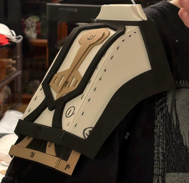 May include: A white foam shoulder armor piece with black trim and brown accents. The armor has a geometric design with a keyhole shape in the center. The armor is partially assembled and has markings on the surface.