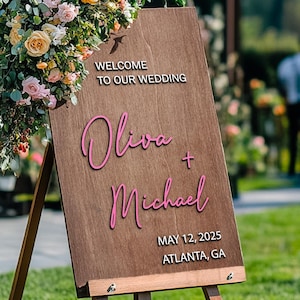 Può includere: Un cartello di legno per matrimonio con un arrangiamento floreale dietro. Il cartello dice "Benvenuti al nostro matrimonio" e "Olivia + Michael" in corsivo rosa. La data "12 maggio 2023" e la località "Atlanta, GA" sono anche sul cartello.