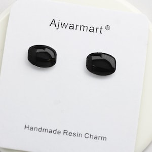 Könnte beinhalten: Ein Paar schwarze Ohrstecker aus Harz in ovaler Form auf einer weißen Karte. Die Ohrringe sind glänzend. Auf der Karte steht "Ajwarmart" und "Handmade Resin Charm".