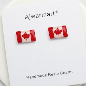 May include: A pair of small stud earrings featuring the Canadian flag design. Each earring has a red border with a white center and a red maple leaf. The earrings are displayed on a white card with the text "Ajwarmart" and "Handmade Resin Charm".
