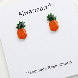 May include: Two small, orange and green pineapple-shaped earrings on a white card. The earrings are handmade resin charms. The card has the text "Ajwarmart" and "Handmade Resin Charm".