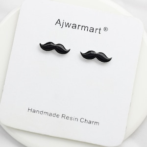 May include: A pair of black mustache-shaped resin charms on a white card. The card has the text "Ajwarmart" at the top and "Handmade Resin Charm" at the bottom. The charms are small and have a glossy finish.