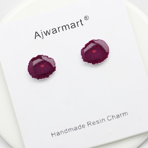 May include: A pair of resin stud earrings. Each earring is a dark burgundy color with a floral design and a small red heart in the center. The earrings are on a white card with the text "Ajwarmart" and "Handmade Resin Charm".