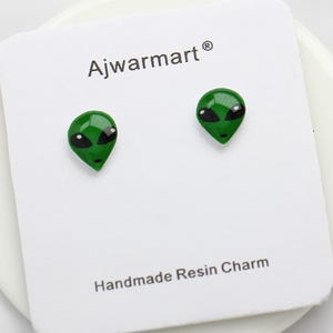Può includere: Orecchini a bottone a forma di alieno verde con occhi neri, presentati su una carta bianca. La carta riporta la scritta "Ajwarmart" e "Handmade Resin Charm". Un accessorio divertente e originale.