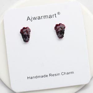 Op de afbeelding: Zwarte schedel-vormige oorstekers met rode en groene details. De oorbellen worden gepresenteerd op een witte kaart met de tekst "Ajwarmart" en "Handmade Resin Charm".