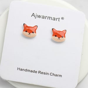 Puede incluir: Pendientes de botón con forma de zorro. Los pendientes son abalorios de resina de color naranja, negro y crema. Se muestran en una tarjeta blanca con el texto "Ajwarmart" y "Handmade Resin Charm".