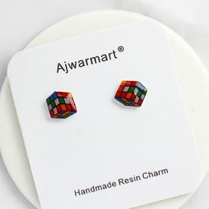 May include: A pair of colorful Rubik's Cube stud earrings on a white card. The card has the text "Ajwarmart" and "Handmade Resin Charm". The earrings are made of resin and feature the classic cube design with red, blue, green, orange, and white squares.