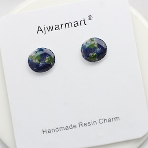 May include: A pair of handmade resin stud earrings featuring a miniature Earth design. The earrings are round, with a blue and green color scheme, and are displayed on a white card with the text "Ajwarmart®" and "Handmade Resin Charm".