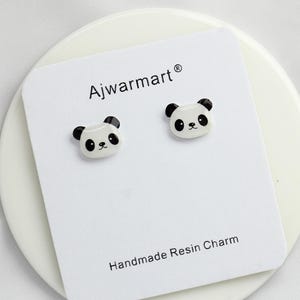 May include: A pair of panda bear stud earrings on a white card. The earrings are white with black accents, featuring a panda face design. The card reads "Ajwarmart®" and "Handmade Resin Charm".