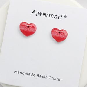 May include: Two red heart-shaped earrings with a flamingo design are displayed on a white card. The card has the text "Ajwarmart" and "Handmade Resin Charm". The earrings are small and have a glossy finish.
