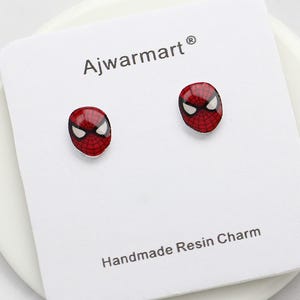 Puede incluir: Un par de pendientes de botón con la cara de Spider-Man en rojo y negro. Los pendientes están sobre una tarjeta blanca con el texto "Ajwarmart" y "Handmade Resin Charm". Los pendientes tienen un acabado brillante.