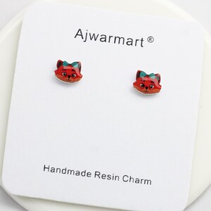 Puede incluir: Un par de pendientes de botón con forma de zorro. Cada pendiente presenta una cara de zorro rojo con un lazo verde sobre un fondo blanco. Los pendientes están sobre una tarjeta blanca con el texto "Ajwarmart" y "Handmade Resin Charm".