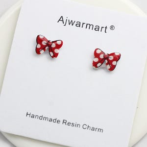 May include: Red bow-shaped stud earrings with white polka dots, displayed on a white card. The card reads "Ajwarmart" and "Handmade Resin Charm". The earrings are a cute accessory.