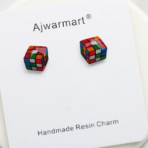 May include: A pair of colorful Rubik's Cube earrings on a white card. The earrings are miniature replicas of the classic puzzle, featuring red, orange, blue, green, yellow, and white squares. The card reads "Ajwarmart" and "Handmade Resin Charm".