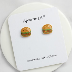 May include: A pair of burger-shaped earrings on a white card. The earrings are small, with a smiling face on each. The card reads "Ajwarmart" and "Handmade Resin Charm". The earrings are a fun, food-themed accessory.