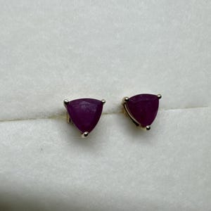May include: A pair of stud earrings featuring triangular-cut deep pinkish-red gemstones set in a metallic prong setting. The stones have a faceted surface that reflects light, showcasing a vibrant color and a distinct three-sided geometric shape.
