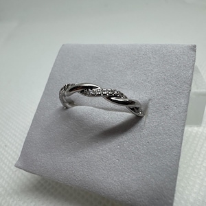 Puede incluir: Un anillo plateado con un diseño trenzado. El anillo está adornado con pequeñas piedras preciosas transparentes engastadas a lo largo de la parte superior de la trenza. El anillo se muestra sobre una superficie gris texturizada.