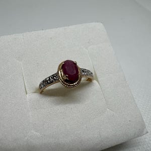 May include: A gold ring featuring a large oval ruby gemstone. The ring has a decorative gold setting around the ruby and small accent stones on the band. The ring is displayed on a white surface.