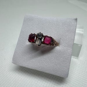 Pode incluir: Um anel dourado com um conjunto de pedras preciosas. O anel apresenta uma disposição central de uma pedra cinza, branca e rosa de corte quadrado, ladeada por duas pedras vermelhas. O anel é exibido em uma superfície branca.