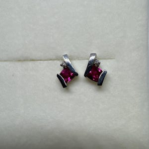 May include: A pair of silver-toned stud earrings featuring a square-cut, vibrant pink gemstone set within a curved metal frame. Each earring is accented with a single, small, clear round stone positioned above the pink square gem.