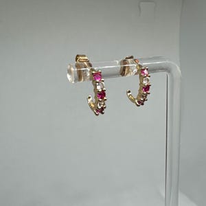 May include: A pair of gold-toned hoop earrings. Each earring features a row of alternating pink and clear gemstones. The earrings are displayed on a clear stand against a light gray background. The gemstones are small and round.