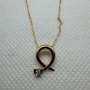 May include: A gold-colored necklace with a ribbon-shaped pendant. The pendant has a small heart-shaped charm with a cluster of small, sparkling stones. The necklace is displayed on a white surface.