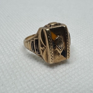May include: A gold-colored ring with a rectangular face featuring a crest design with black, yellow, and white accents. The ring has the year "1919" on the side. The ring is likely a class ring or a commemorative piece.