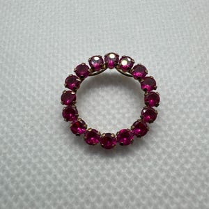 May include: A circular brooch featuring a ring of vibrant, round, ruby-red gemstones set in a gold-toned metal. The gemstones are evenly spaced around the circle, creating a continuous, sparkling design. The brooch is set against a white background.