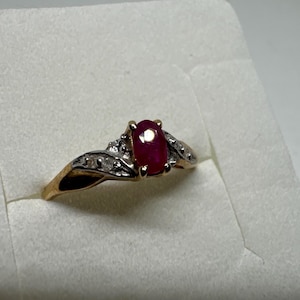 Peut inclure: Une bague en or avec une pierre centrale en forme de rubis ovale. La bague a un design à tige fendue avec de petits diamants de chaque côté du rubis. La bague est posée sur une surface blanche.