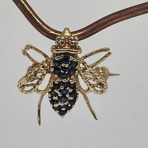 Puede incluir: Un broche de abeja de oro con un diseño detallado. El cuerpo de la abeja está cubierto de pequeños zafiros azules y las alas están hechas de un trabajo de oro intrincado.