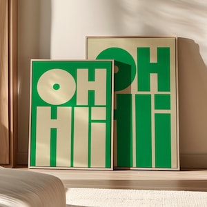 May include: Two framed art prints with the words "OH Hi!" in a bold, green font against a cream background. The prints are rectangular and have gold frames. The artwork is displayed on a light-colored wall, with natural light and shadows.