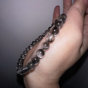 May include: A beaded bracelet featuring translucent, smoky quartz spheres. The beads have internal inclusions, creating a shimmering effect. The bracelet is strung on a clear elastic cord, and is a neutral accessory.