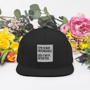 May include: Black flat-brimmed baseball cap with the text "TIME IS NON REFUNDABLE. USE IT WITH INTENTION." in a white square. The cap is surrounded by colorful flowers, including pink peonies and yellow daffodils.