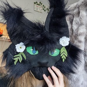 May include: A black faux fur cat mask featuring tall, pointed ears with tufted tips, bright green eyes with blue pupils, and two white fabric flowers accented with small green leaf charms attached to the sides of the face.