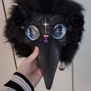 May include: A black plague doctor mask with a long beak, adorned with black fur, roses, and gemstones. The mask features large, detailed eyes with blue and white accents, and a small silver bell at the beak's tip. A hand holds the mask.