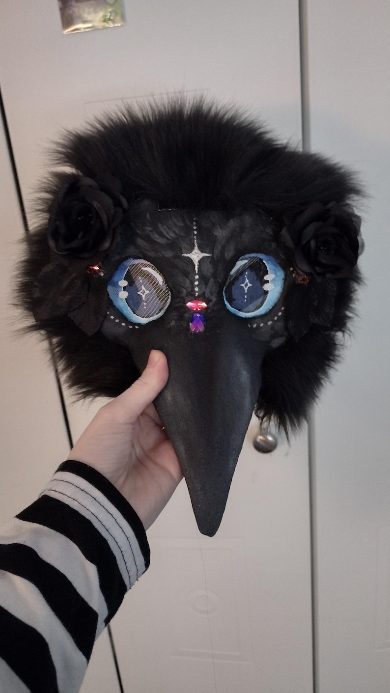 May include: A black plague doctor mask with a long beak, adorned with black fur, roses, and gemstones. The mask features large, detailed eyes with blue and white accents, and a small silver bell at the beak's tip. A hand holds the mask.