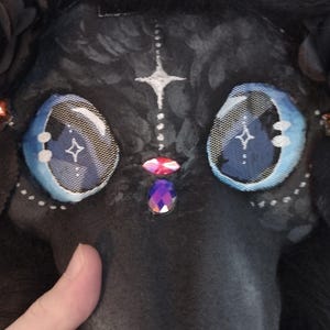May include: A black plague doctor mask with large, detailed blue eyes and a long beak. The mask is adorned with white accents, a star design, and decorative jewels. The top of the mask is covered in black fur.