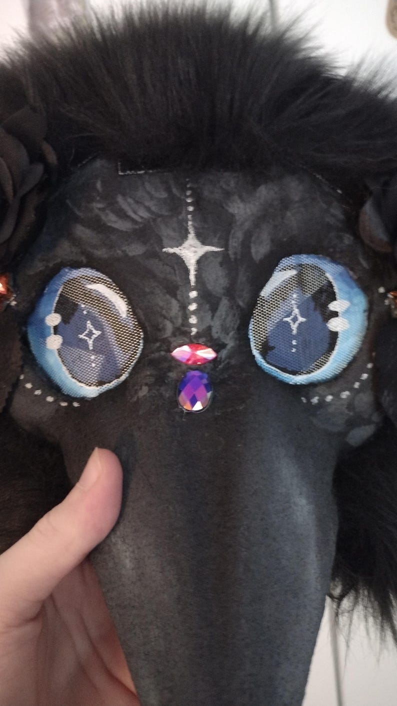 May include: A black plague doctor mask with large, detailed blue eyes and a long beak. The mask is adorned with white accents, a star design, and decorative jewels. The top of the mask is covered in black fur.